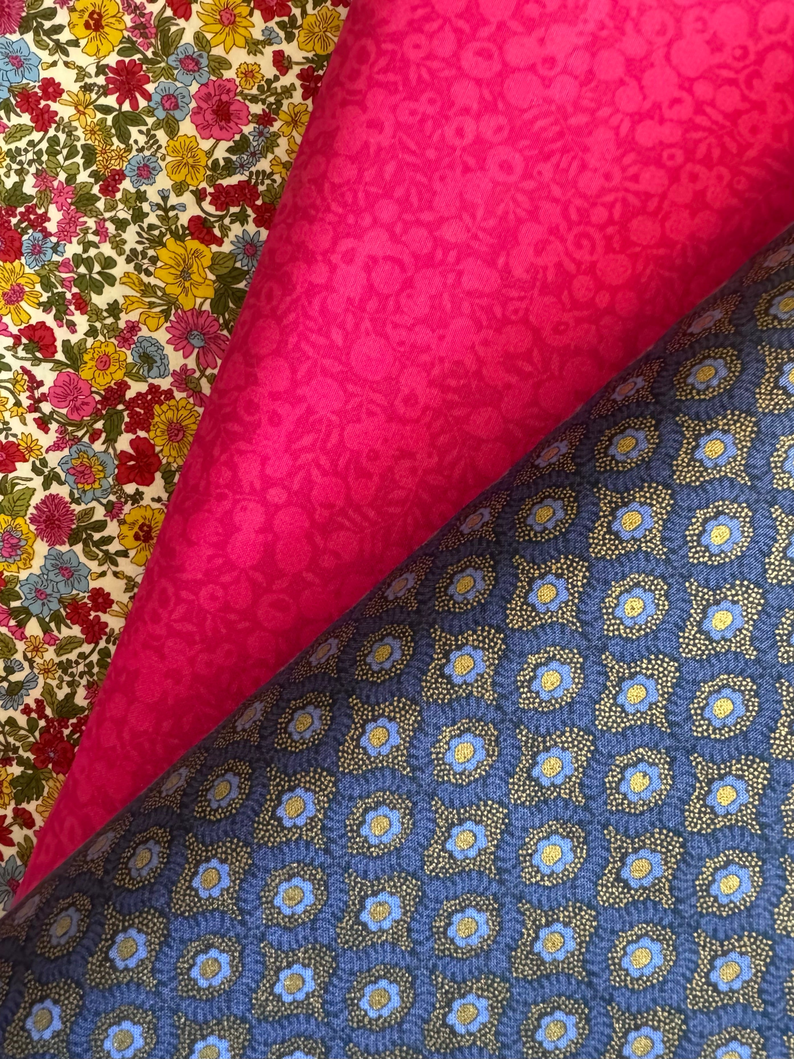 Three folded fabric samples displayed diagonally: a bright pink floral fabric, a multicolor floral fabric with red, yellow, and blue flowers on a cream background, and a blue fabric with repeating gold and light blue floral medallion patterns.
