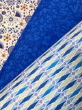 Three folded fabric samples displayed diagonally, showcasing different textile patterns: a cream and blue floral tile pattern, a solid royal blue floral jacquard, and a blue, white, and light purple geometric diamond pattern.