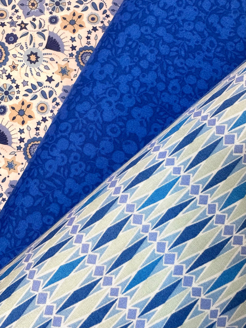 Three folded fabric samples displayed diagonally, showcasing different textile patterns: a cream and blue floral tile pattern, a solid royal blue floral jacquard, and a blue, white, and light purple geometric diamond pattern.
