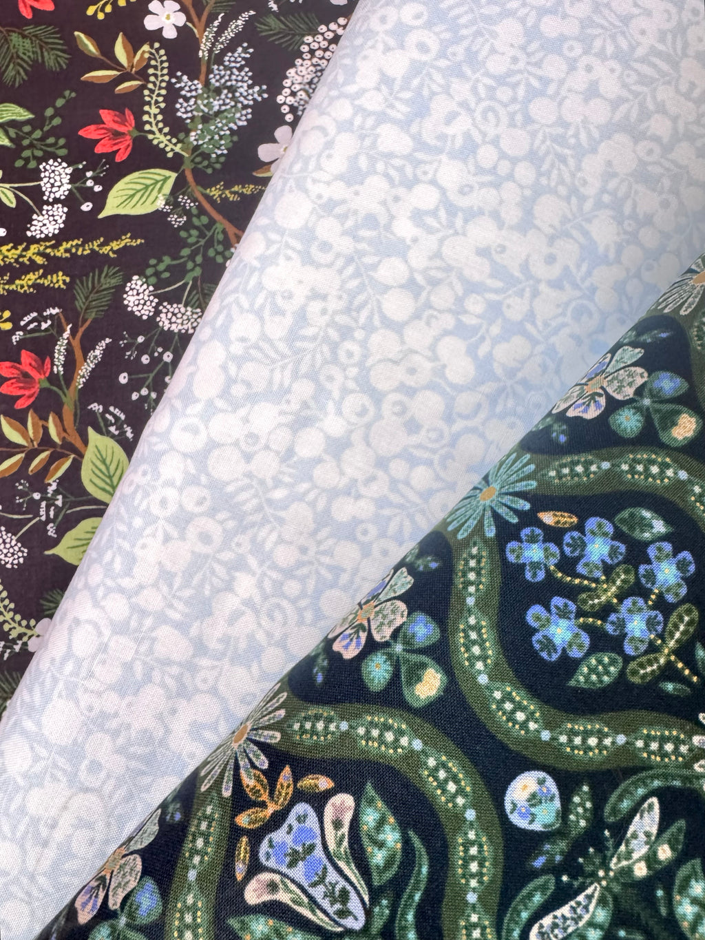 Three rolls of patterned fabric laid diagonally across the frame. The top-left roll has a dark floral print with red, green, and white botanical motifs on a black background.