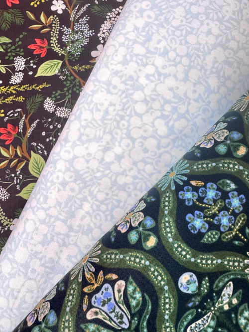 Three rolls of patterned fabric laid diagonally across the frame. The top-left roll has a dark floral print with red, green, and white botanical motifs on a black background.
