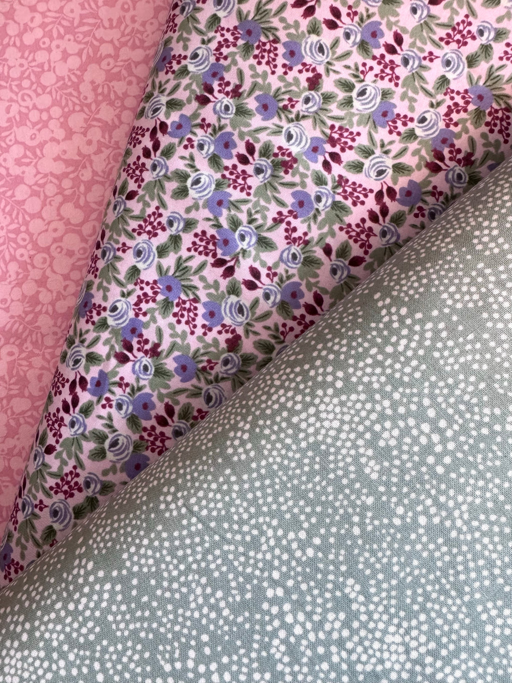 Three folded fabric samples displayed diagonally, showing different patterns: a pink damask pattern, a light pink floral print with purple and blue flowers, and a sage green fabric with white polka dots.