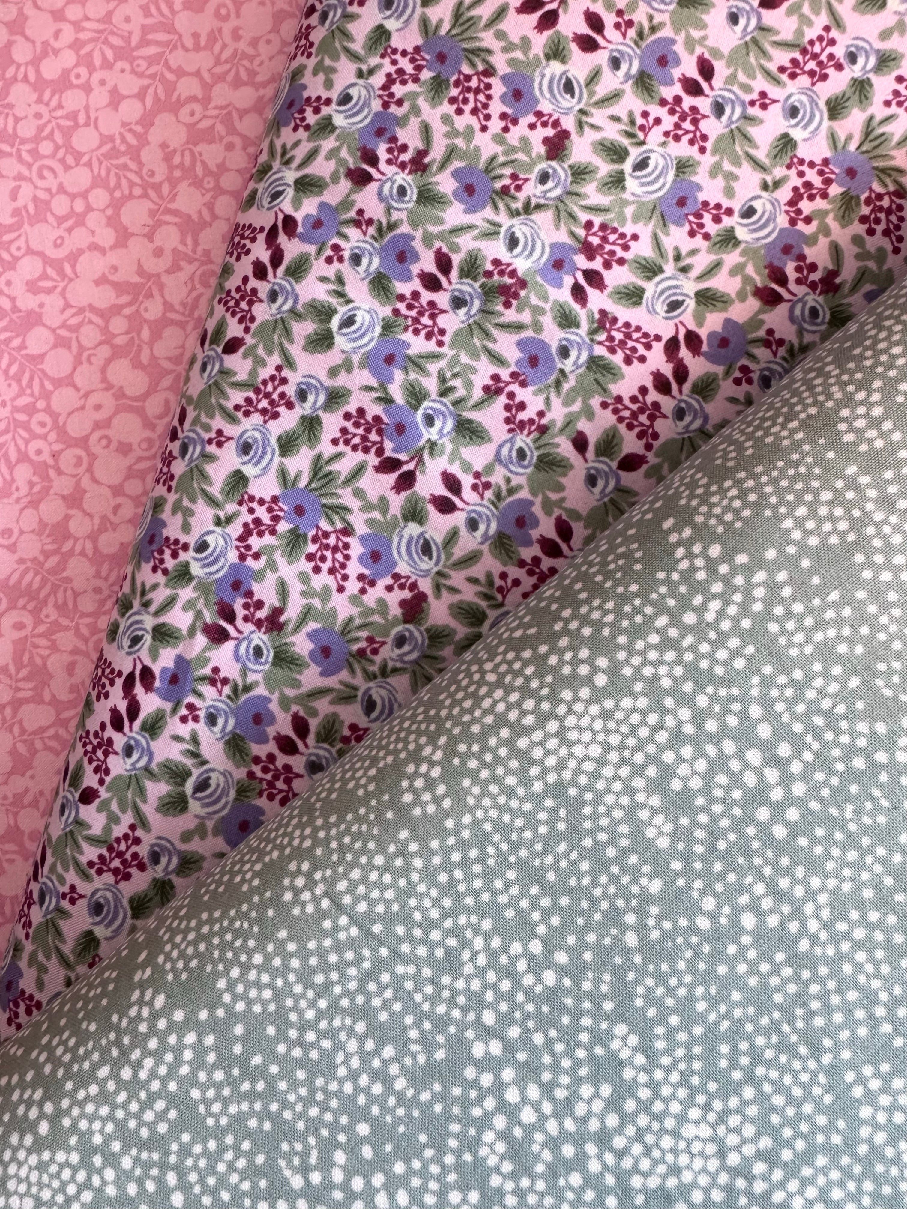 Three folded fabric samples displayed diagonally, showing different patterns: a pink damask pattern, a light pink floral print with purple and blue flowers, and a sage green fabric with white polka dots.