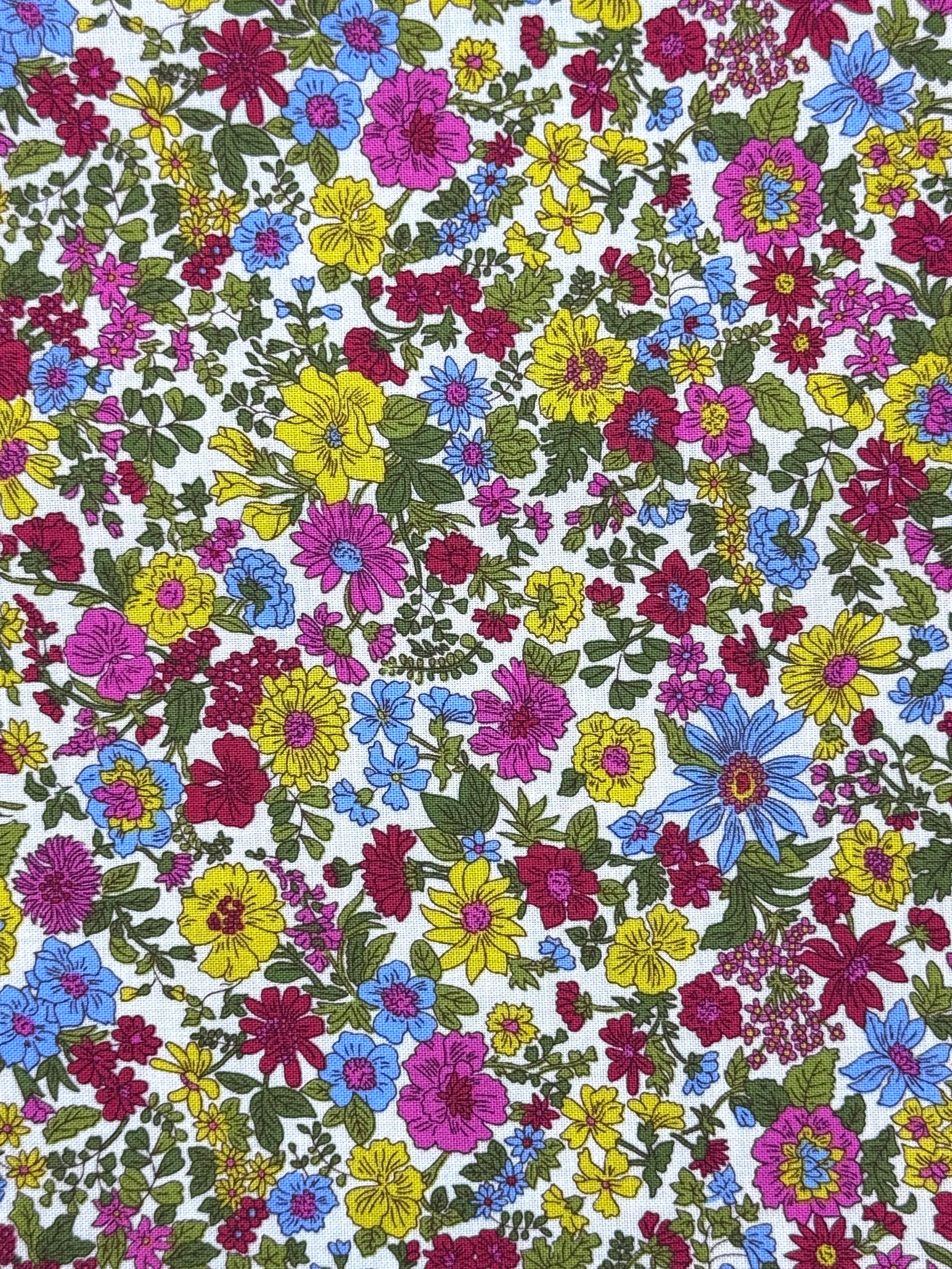 A close-up of a white fabric with a dense, colorful floral print featuring yellow, pink, blue, and red flowers with green leaves.