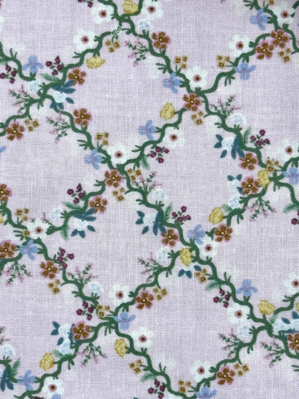 A close-up of a light lavender fabric with a repeating embroidered floral vine pattern in green, white, yellow, blue, and purple.