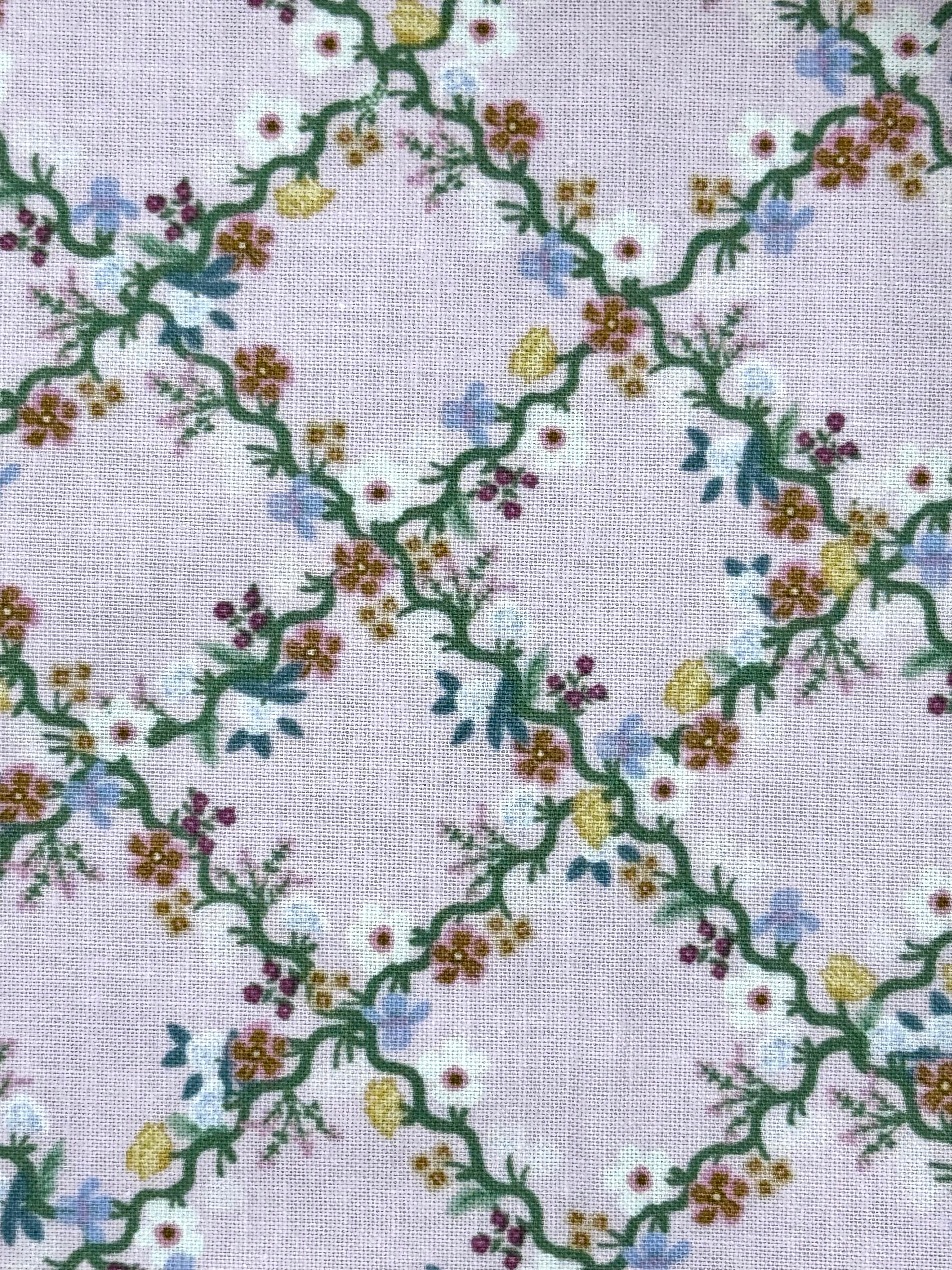 A close-up of a light lavender fabric with a repeating embroidered floral vine pattern in green, white, yellow, blue, and purple.