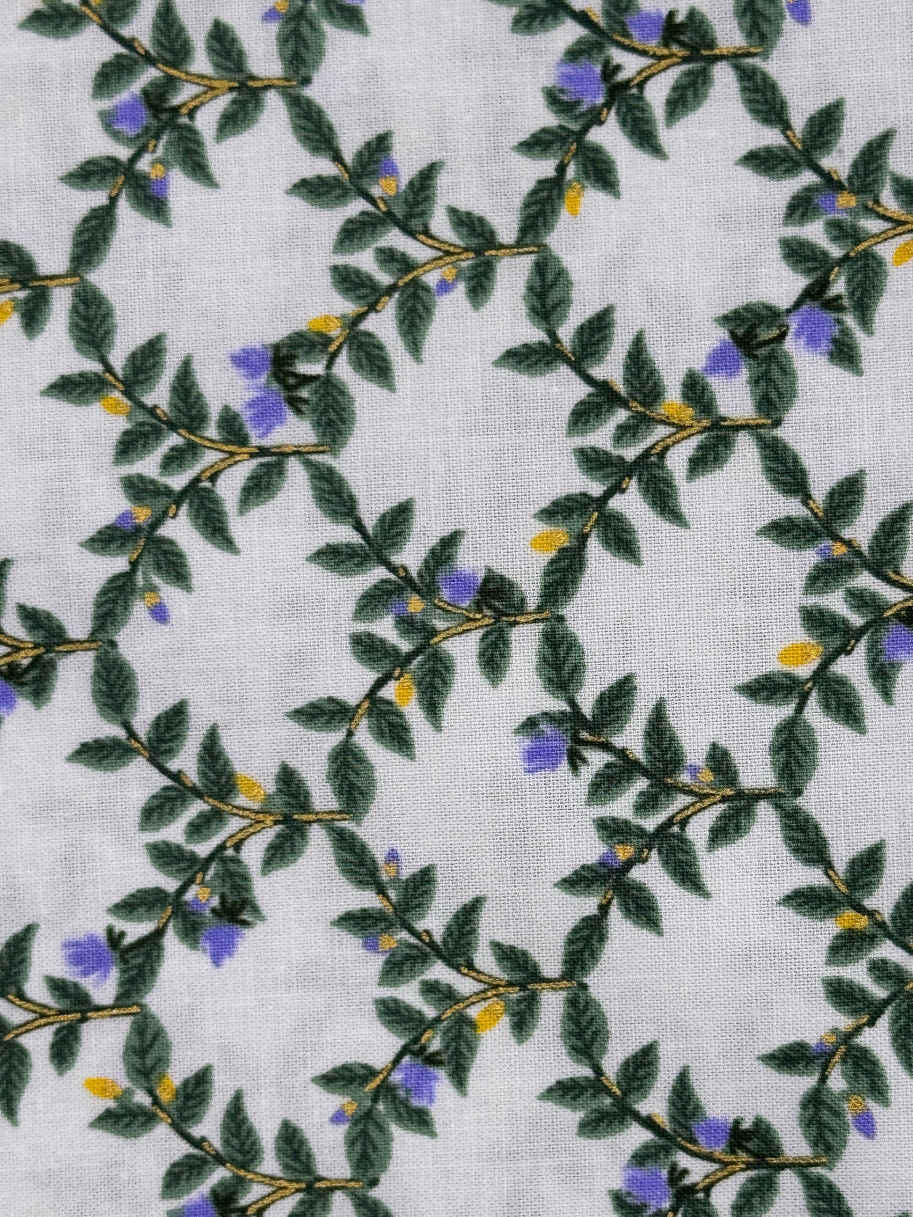 A close-up of a white textured fabric featuring a repeating botanical pattern of green leaves, thin brown branches, and small purple and yellow flowers.