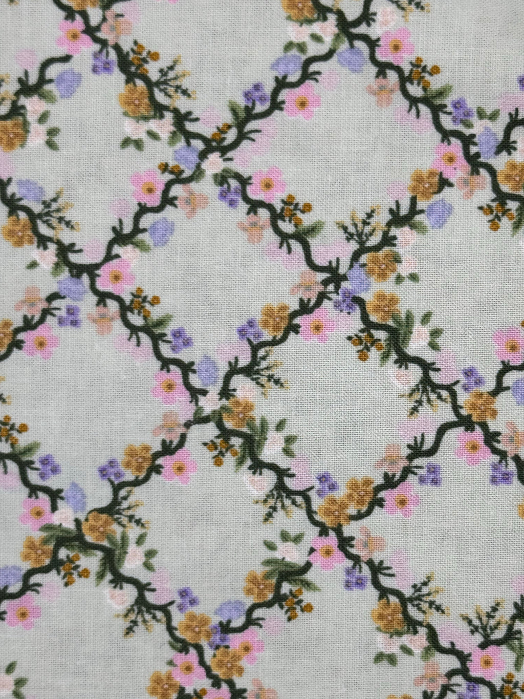 A close-up of a fabric pattern featuring a trellis design with dark green vines and small flowers in pink, purple, yellow, and orange on a light gray background.
