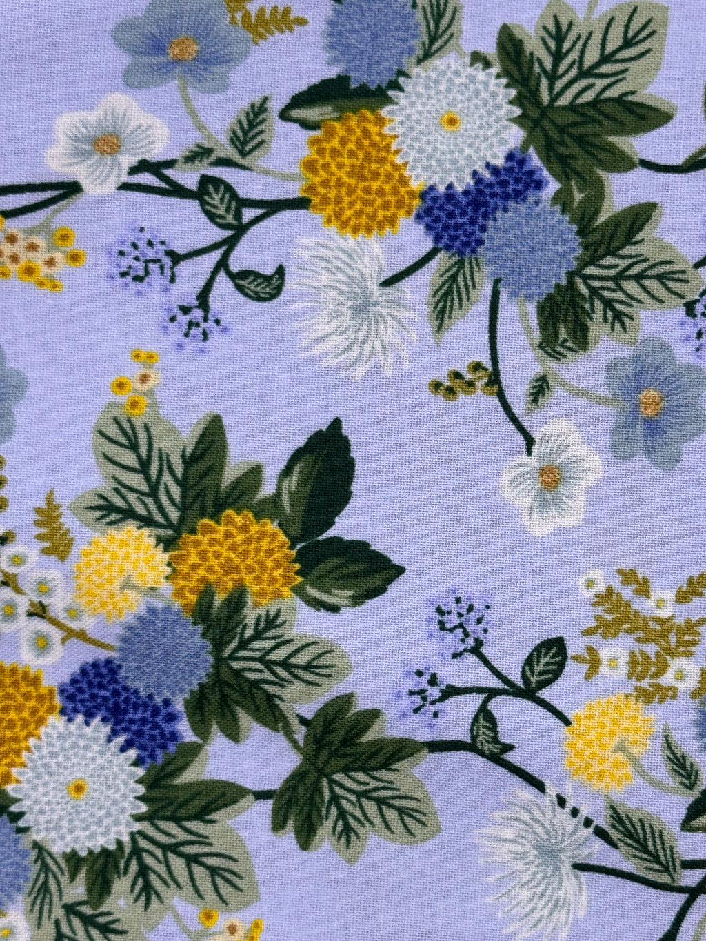 A close-up of a floral fabric pattern on a light lavender background, featuring white, blue, and yellow flowers with green leaves and stems.