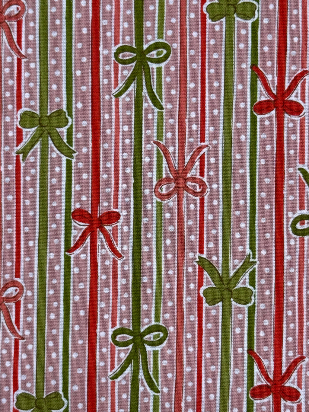 A close-up of a patterned fabric featuring vertical red and green stripes with white polka dots, interspersed with green and red ribbon-bow motifs.