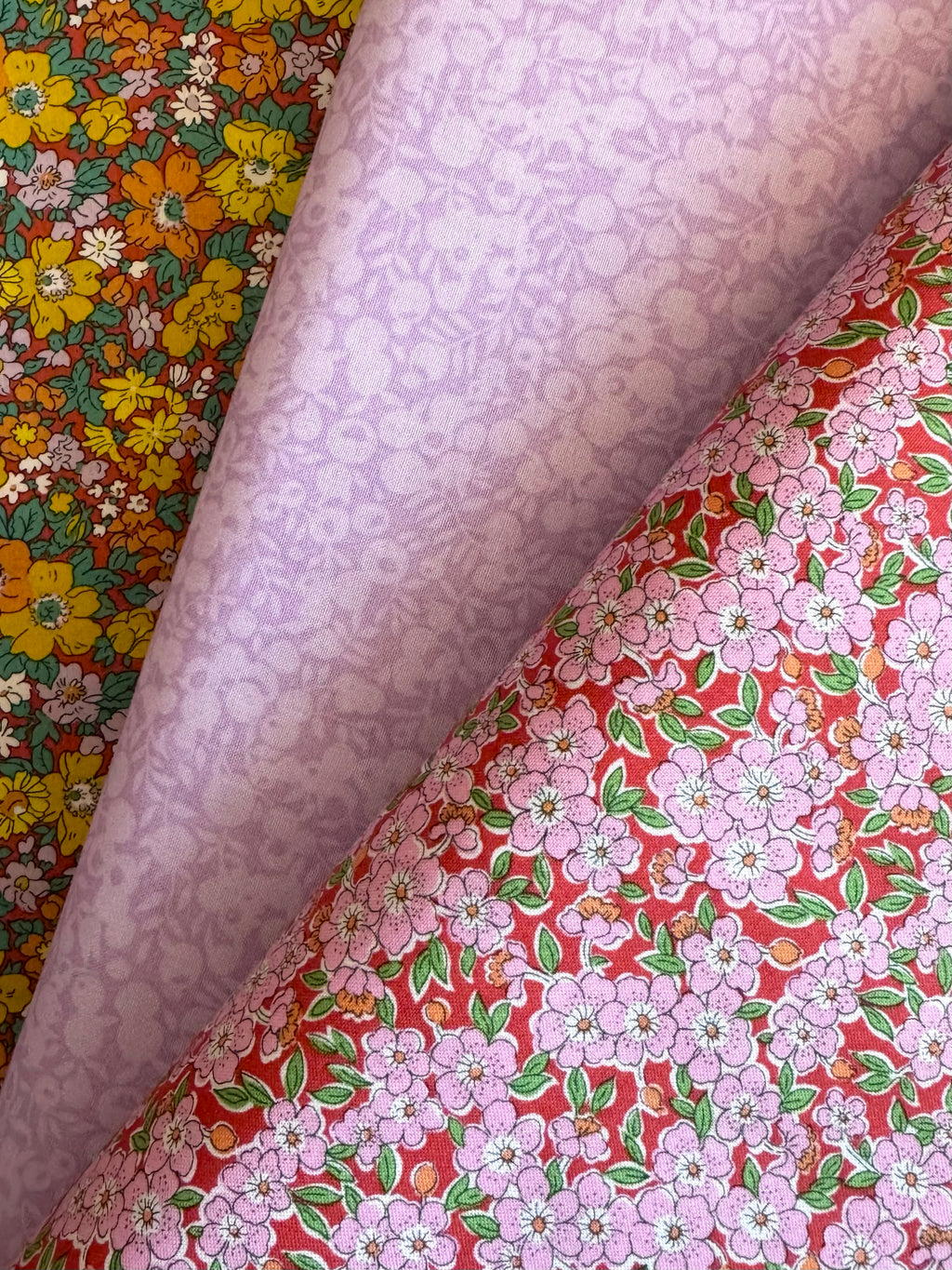 Three rolls of patterned fabric stacked diagonally, showing different designs: a yellow and orange floral print, a light lavender floral print, and a red floral print with pink blossoms and green leaves.