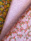 Three rolls of patterned fabric stacked diagonally, showing different designs: a yellow and orange floral print, a light lavender floral print, and a red floral print with pink blossoms and green leaves.