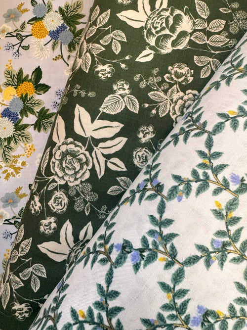 Three folded fabric samples with floral and botanical patterns, arranged diagonally. The top-left fabric has multicolor flowers on a light background.