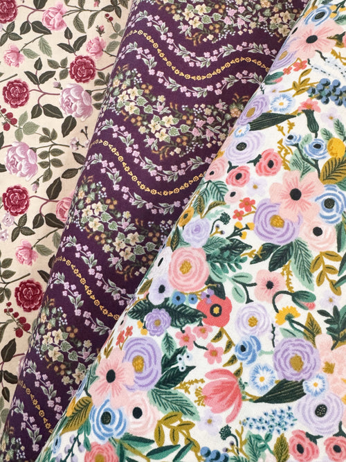 Three folded fabric rolls with floral patterns, showing a cream rose print, a deep purple floral print with gold chain details, and a white floral print with pink, purple, blue, and green flowers.