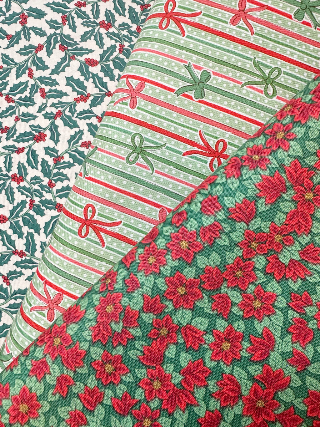 Three Christmas-themed patterned fabrics arranged diagonally, featuring a holly and berry print, a striped design with bows, and a red poinsettia floral pattern on green.